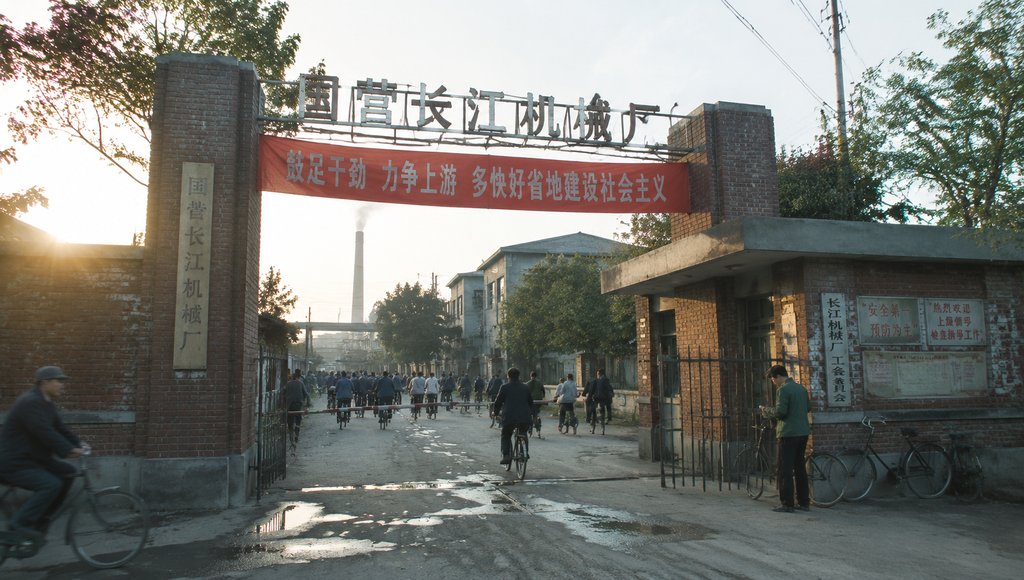 国营长江机械厂大门,红色标语横幅,职工骑自行车进出,年代怀旧写实场景