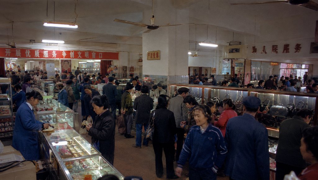 上世纪百货大楼内景，玻璃柜台陈列商品，营业员与顾客互动，吊扇悬挂，怀旧写实风格