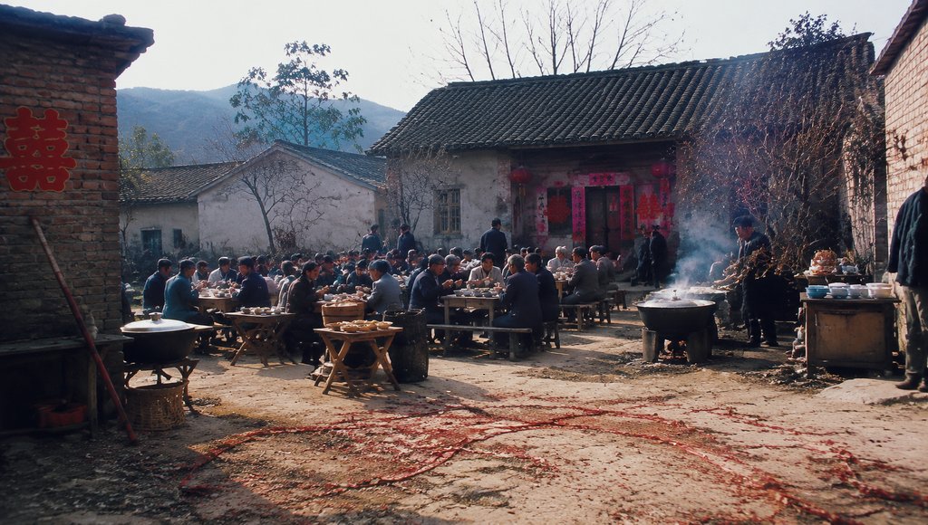农村婚宴场景,八仙桌摆满大锅菜,人群围坐,喜字装饰,年代怀旧写实风格