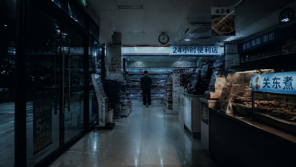 夜晚便利店内部，荧光灯照亮货架与收银台，一人背影伫立，现代都市写实风格