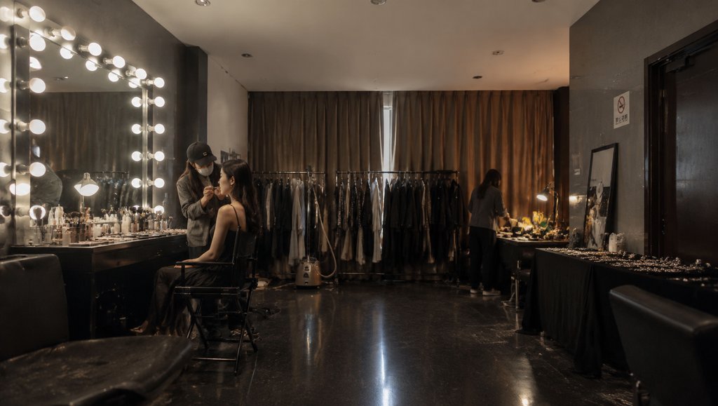 化妆师为模特补妆，镜灯化妆台与服装架环绕，现代都市写实风格