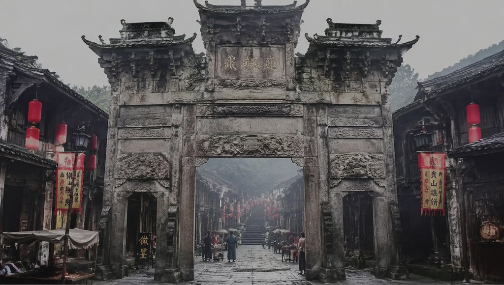 古风古镇石质拱门牌楼，斑驳浮雕与匾额，红灯笼点缀，两侧古建筑林立，雾气弥漫，人物穿行其间